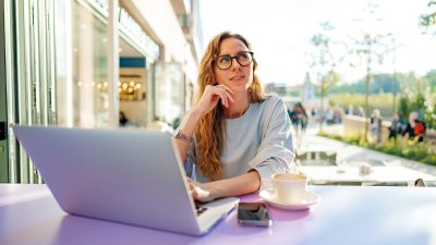 Nainen istuu ulkona kahvilla tietokoneen kanssa. Katsoo mietteliäästi taivasta kohti.