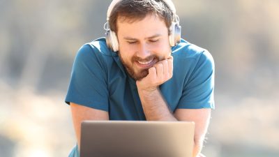 Mies katsoo hymyillen tietokonetta kuulokkeet päässään.