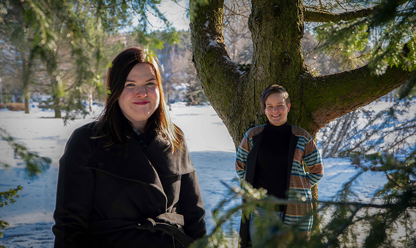 Mirva ja Tanja seisovat hymyillen lumisen maiseman edessä puun alla.