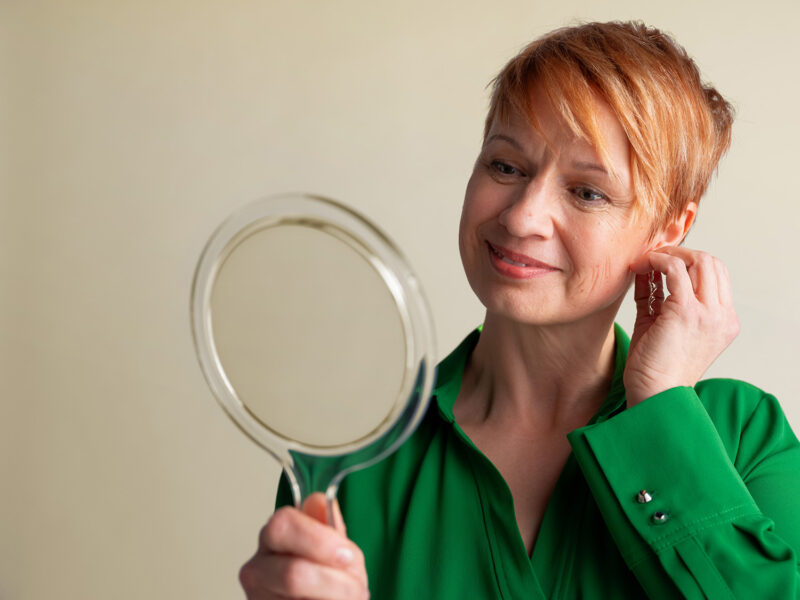 Nainen vihreässä paidassa katsoo peilistä itsevarmana ja asettelee korvista.